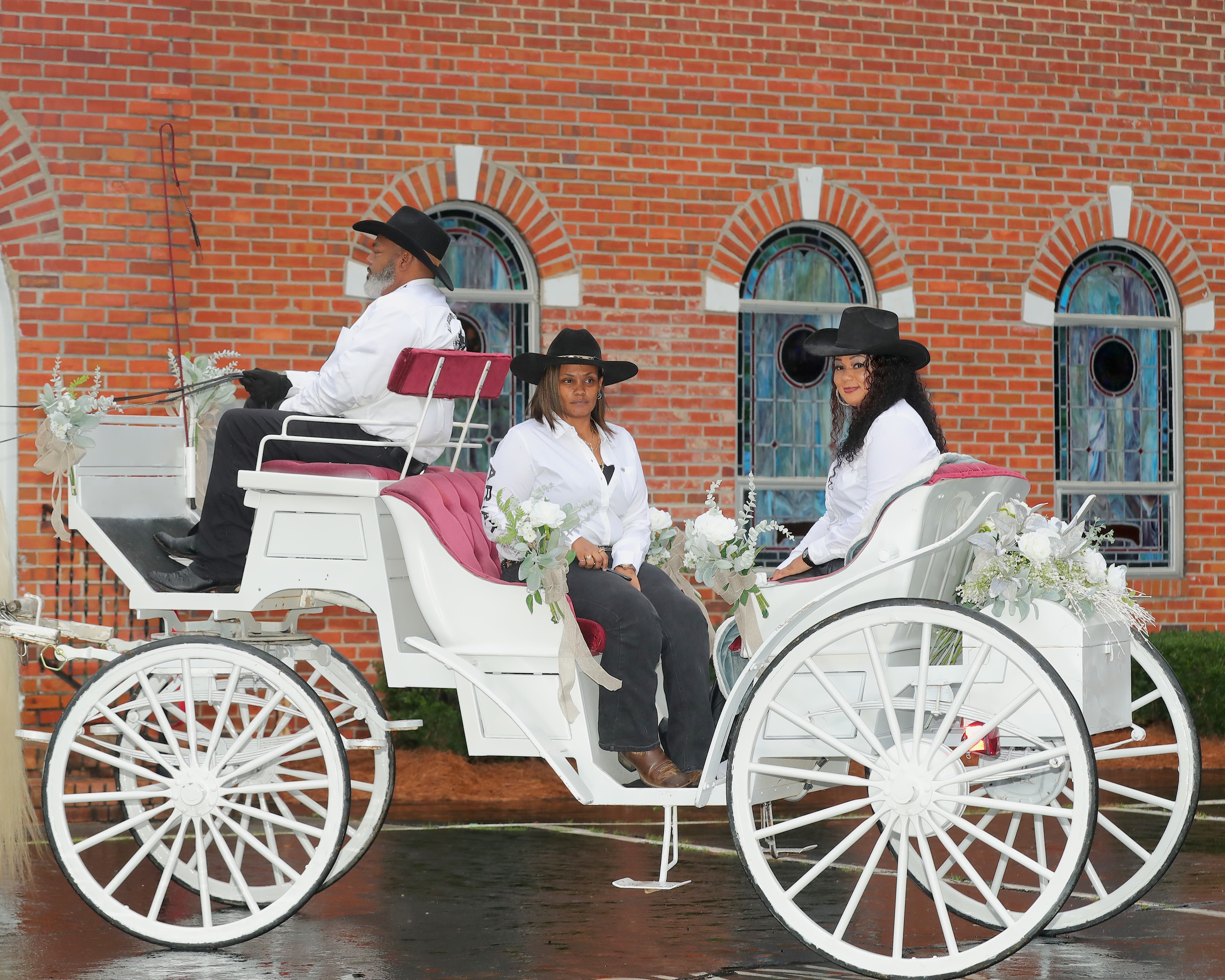 Three people in western attire on a white carriage in front of a brick church